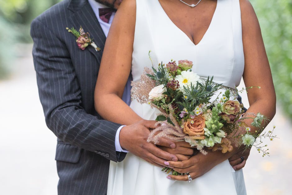 Close-up of a bride and groom holding a floral bouquet arrangement during a wedding, with the bride wearing a white dress and the groom in a dark pinstripe suit. The bouquet features a mix of flowers 